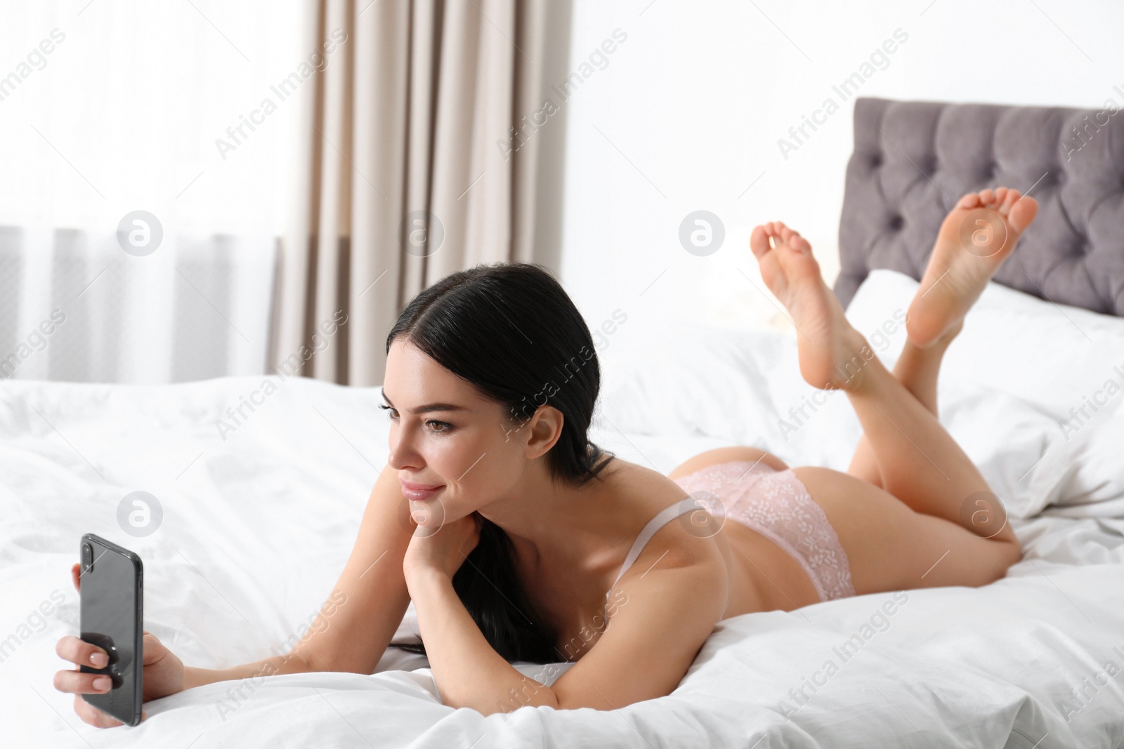 Photo of Beautiful young woman in pink underwear with phone on bed at home