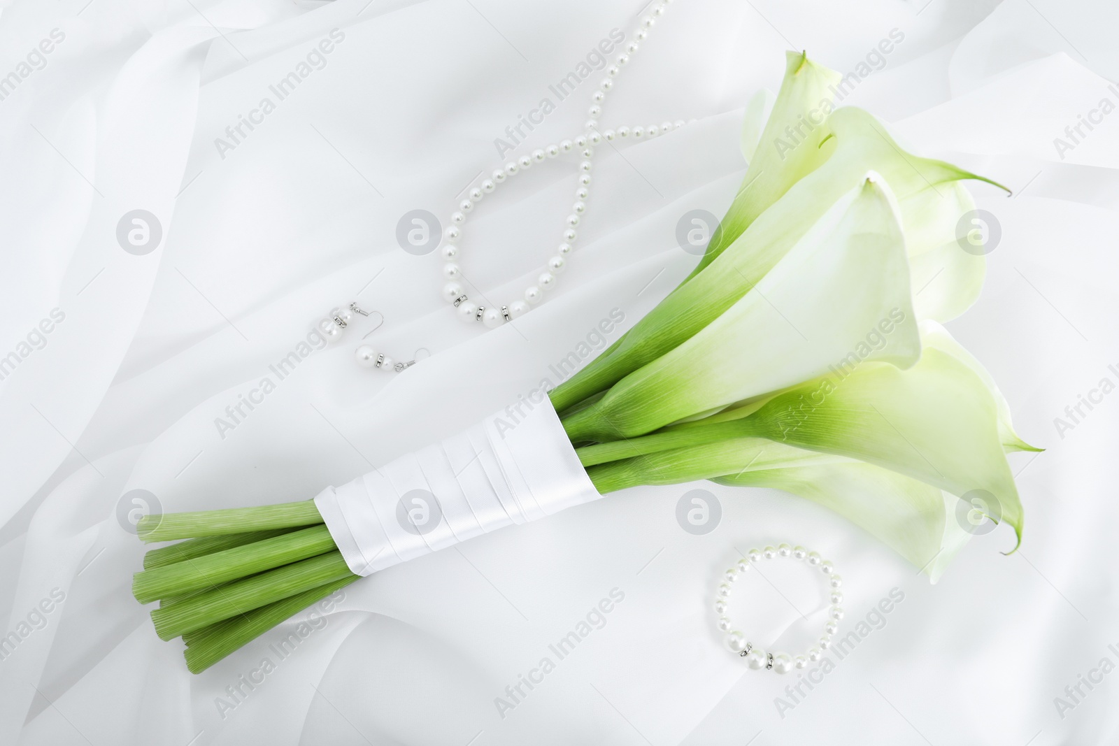 Photo of Beautiful calla lily flowers tied with ribbon and jewelry on white fabric, flat lay