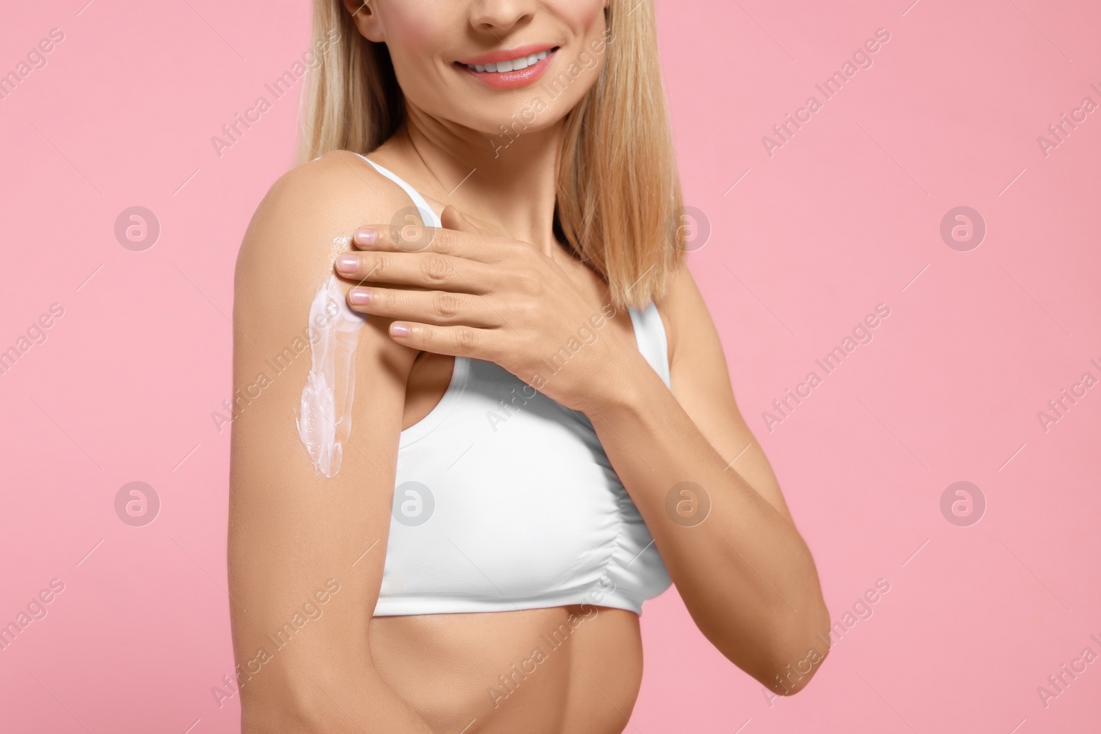 Photo of Woman applying body cream onto her arm against pink background, closeup. Space for text