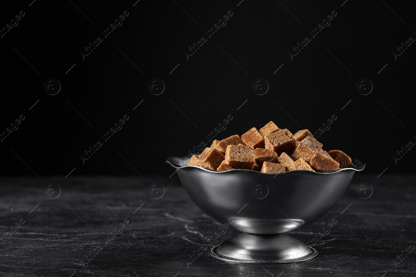 Photo of Metal bowl with brown sugar cubes on black table. Space for text