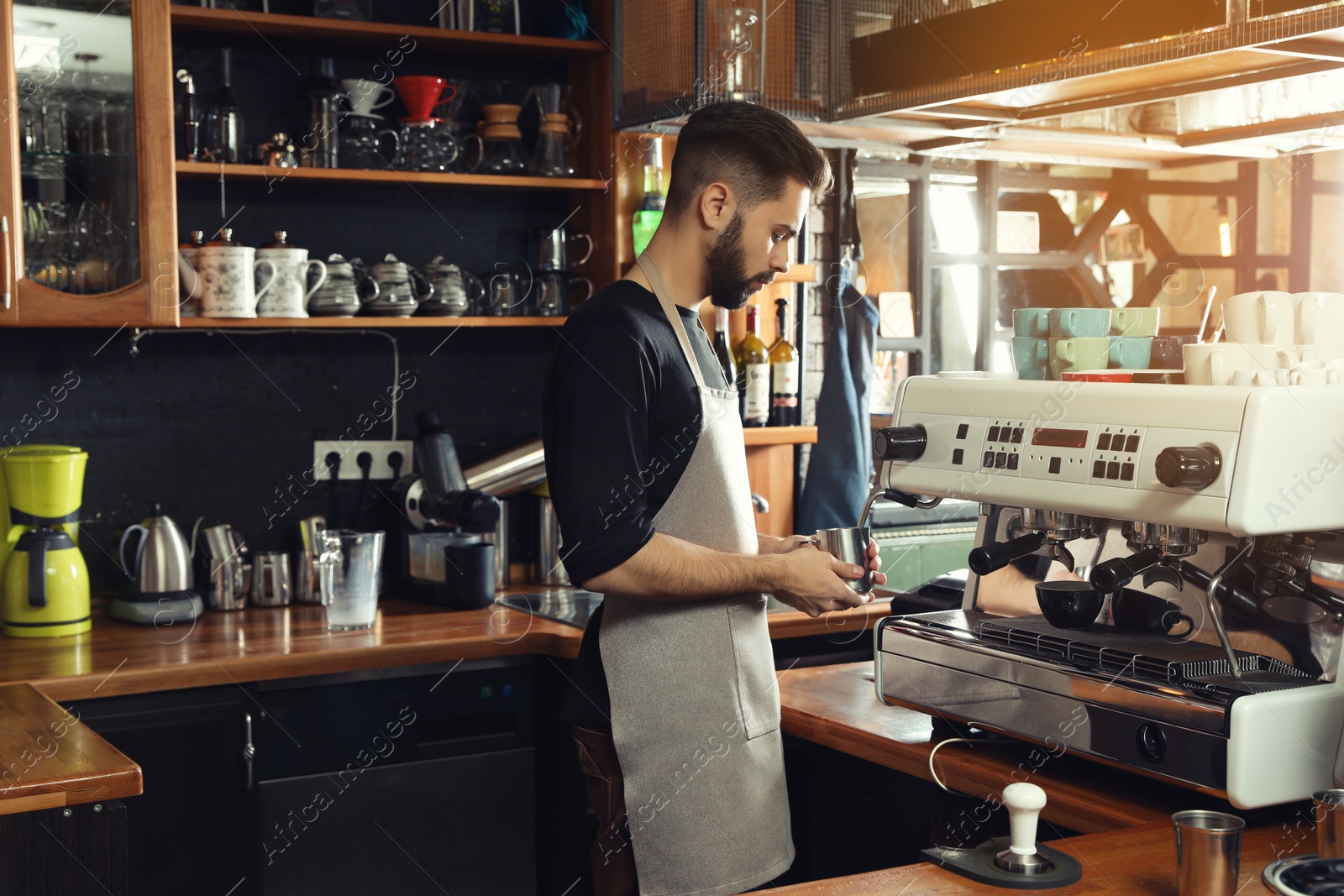 Photo of Barista frothing milk in metal pitcher with coffee machine steam wand at bar counter