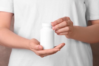 Woman holding blank white bottle with vitamin pills against light brown background, closeup