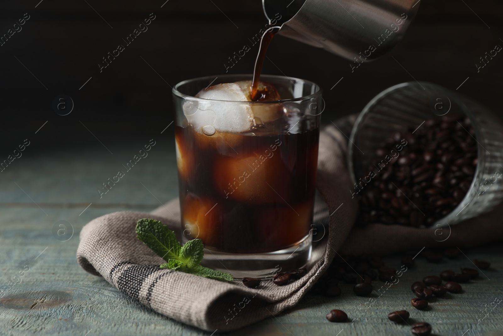 Photo of Pouring coffee into glass with ice cubes, mint and beans on light blue wooden table