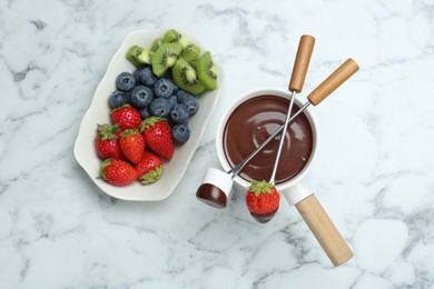 Fondue pot with melted chocolate, sweet marshmallow, fresh kiwi, different berries and forks on white marble table, flat lay