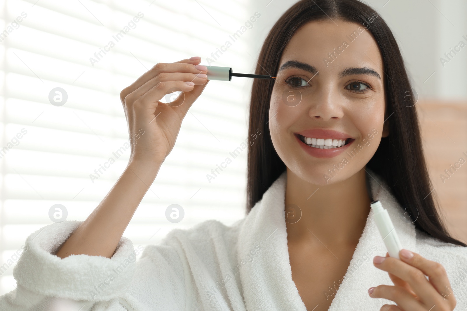 Photo of Beautiful woman applying serum onto her eyelashes indoors, reflection in mirror. Cosmetic product