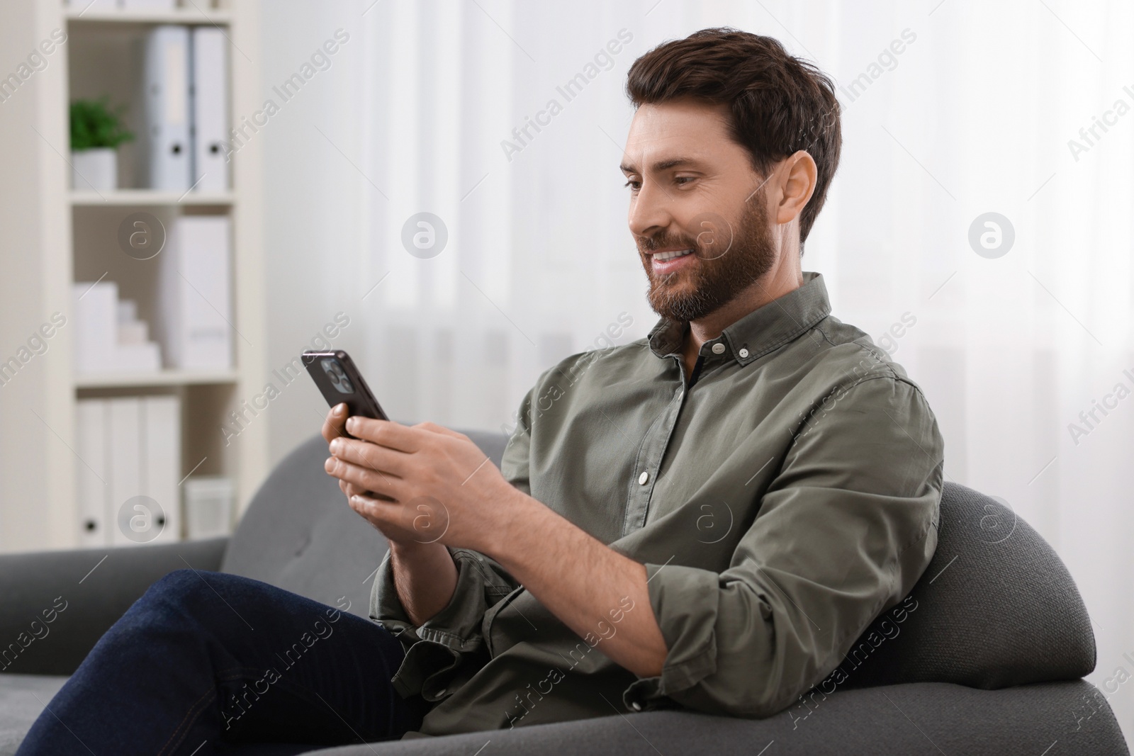 Photo of Smiling man using smartphone on sofa at home