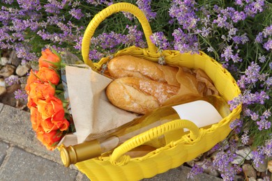 Photo of Yellow wicker bag with beautiful roses, bottle of wine and baguettes near lavender flowers outdoors, above view