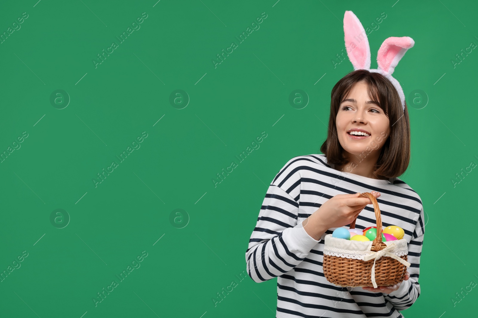 Photo of Easter celebration. Happy woman with bunny ears and wicker basket full of painted eggs on green background, space for text