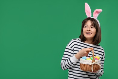 Photo of Easter celebration. Happy woman with bunny ears and wicker basket full of painted eggs on green background, space for text