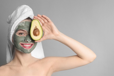 Photo of Young woman with clay mask on her face holding avocado against grey background, space for text. Skin care