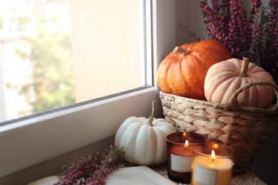 Beautiful heather flowers, burning candles, open book and wicker basket with pumpkins near window indoors. Space for text