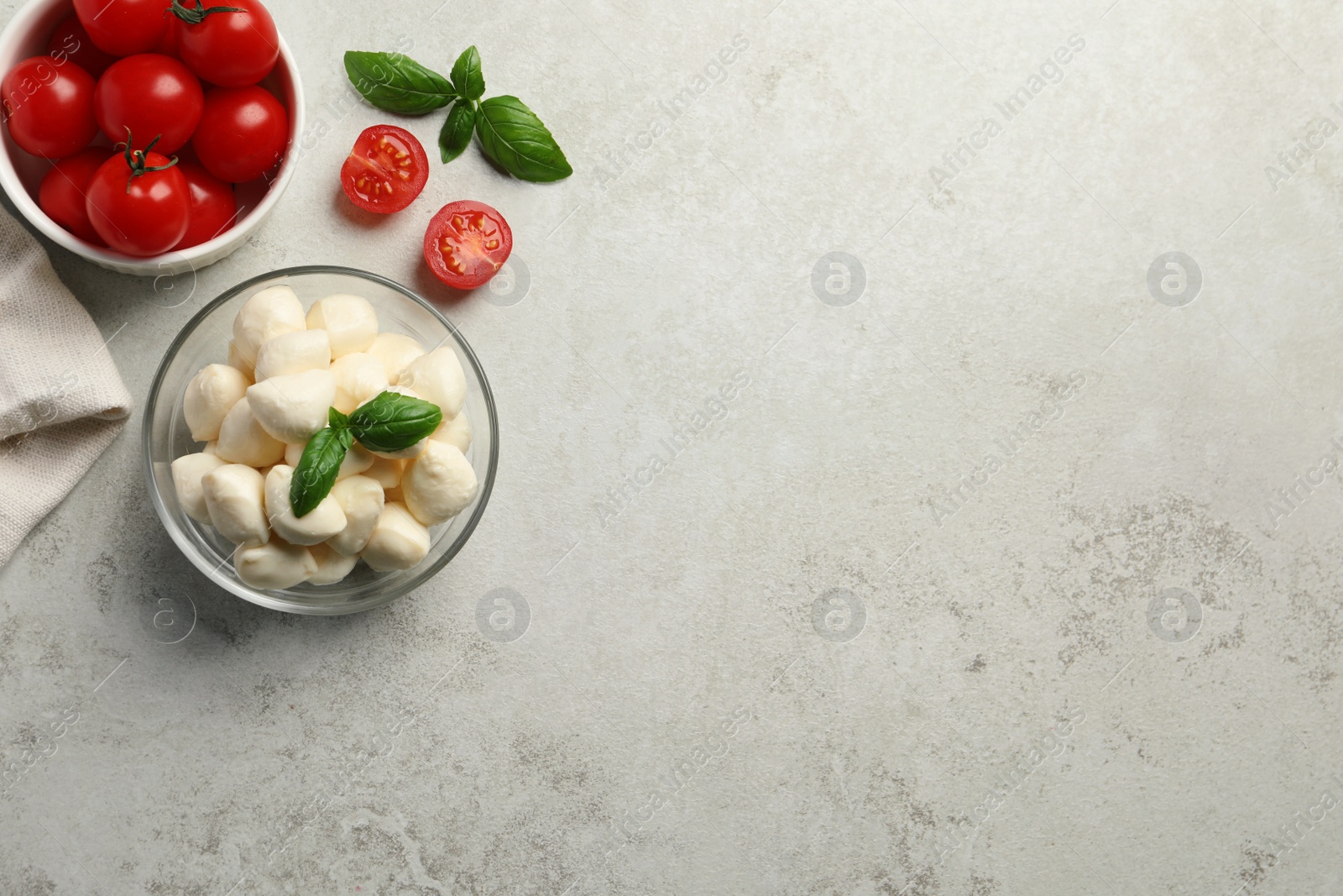 Photo of Delicious mozzarella balls in glass bowl, tomatoes and basil leaves on light gray table, flat lay. Space for text
