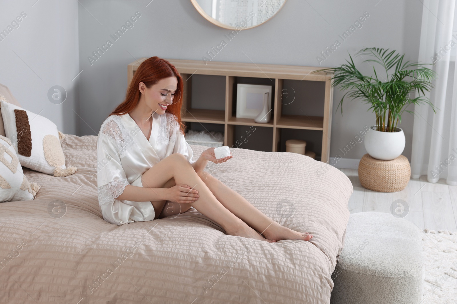 Photo of Beautiful young woman applying body cream onto legs in bedroom, space for text