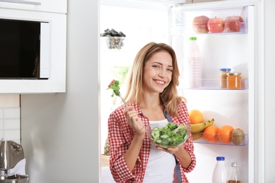 Woman holding bowl with vegetable salad near fridge in kitchen. Healthy diet