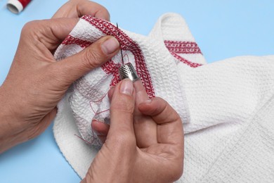 Photo of Woman embroidering on fabric with thimble and sewing needle against light blue background, closeup