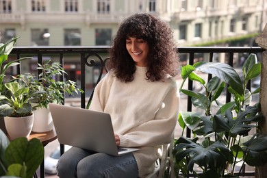 Photo of Beautiful young woman using laptop surrounded by green houseplants on balcony