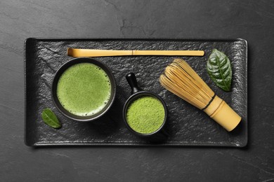 Cup of fresh matcha tea, bamboo whisk, spoon and green powder on black textured table, top view