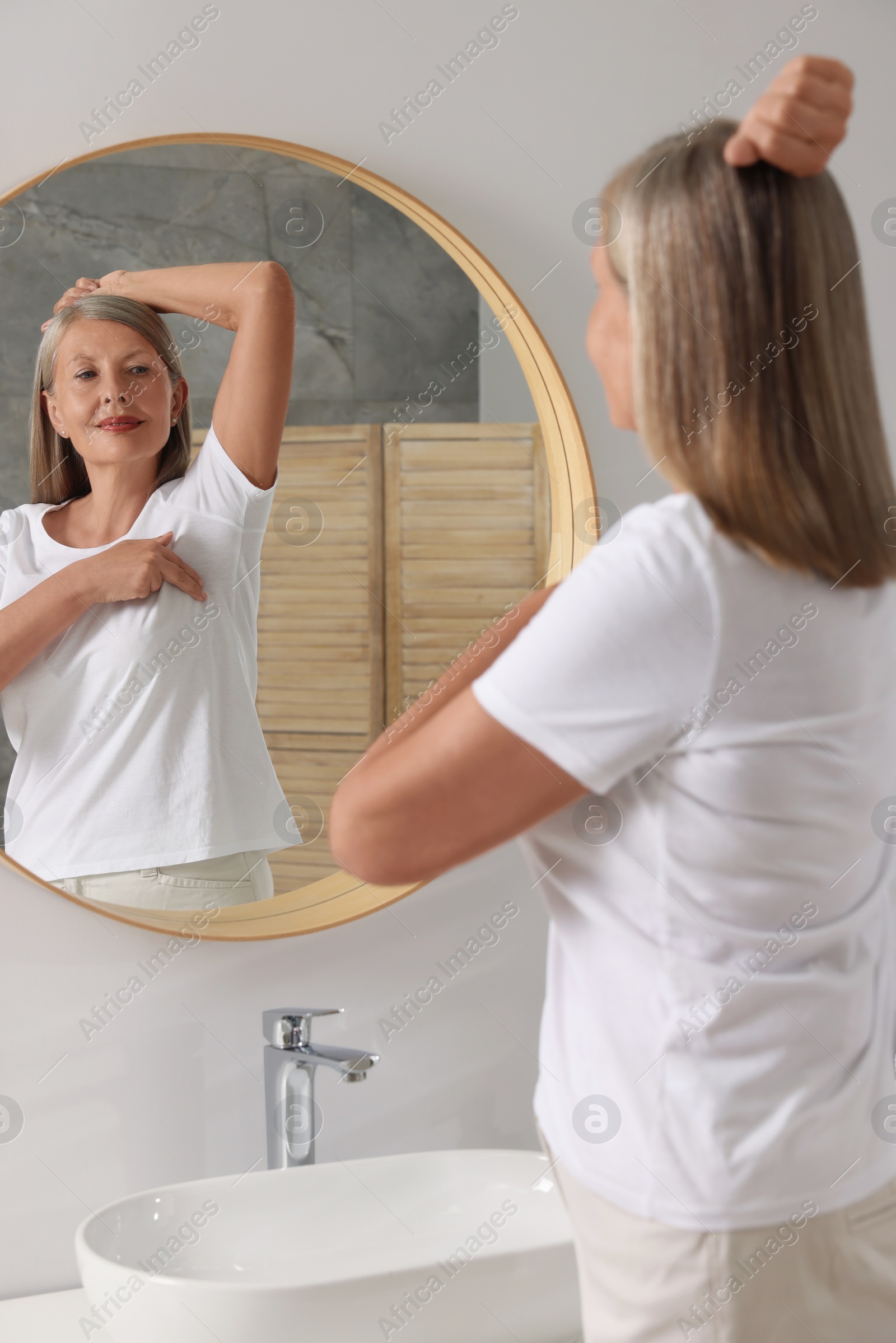 Photo of Beautiful senior woman doing breast self-examination near mirror in bathroom