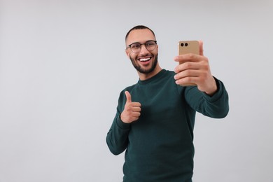 Smiling young man taking selfie with smartphone and showing thumbs up on grey background, space for text