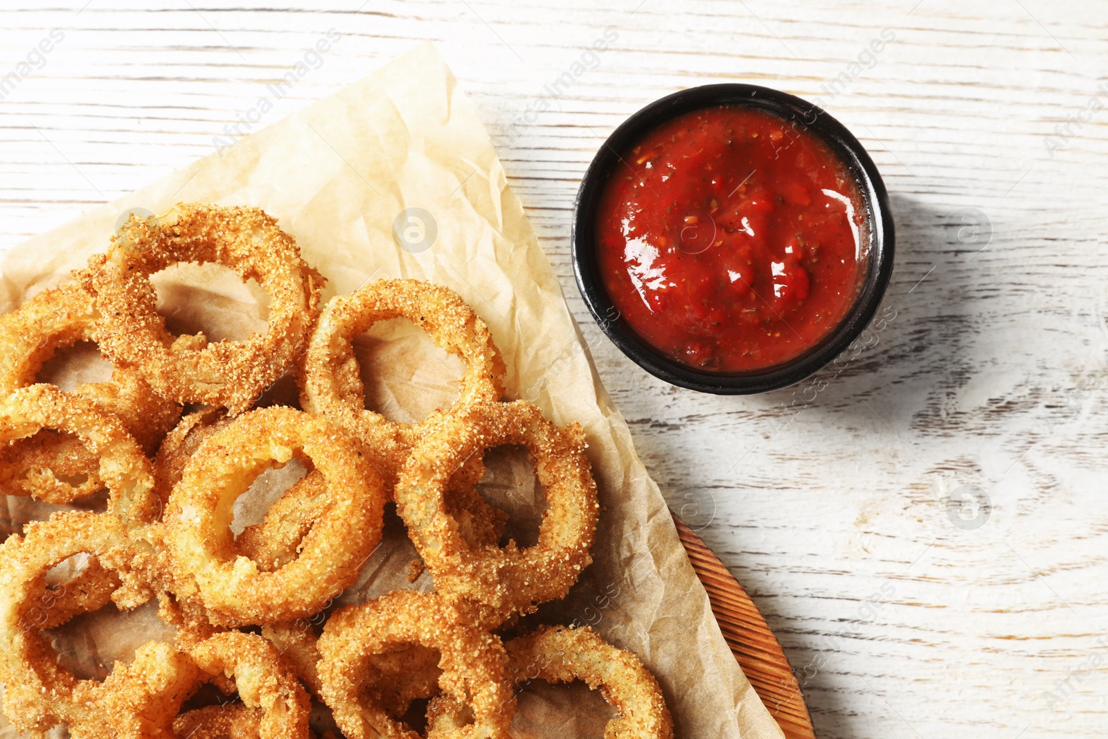 Photo of Homemade crunchy fried onion rings with tomato sauce on wooden table, top view