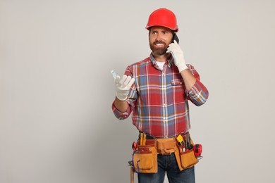 Photo of Professional builder in hard hat with tool belt talking on phone against light background, space for text