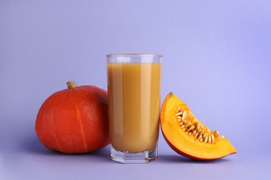 Photo of Tasty pumpkin juice in glass, whole and cut pumpkins on lavender color background