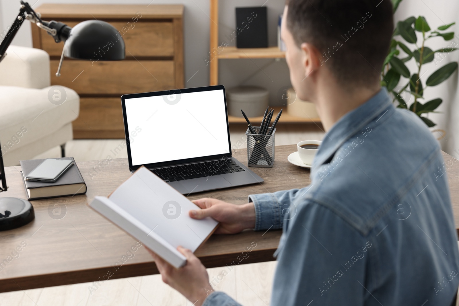 Photo of E-learning. Young man with book during online lesson at table indoors, closeup