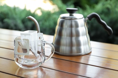 Photo of Glass cup with drip coffee bag and kettle on wooden table, closeup