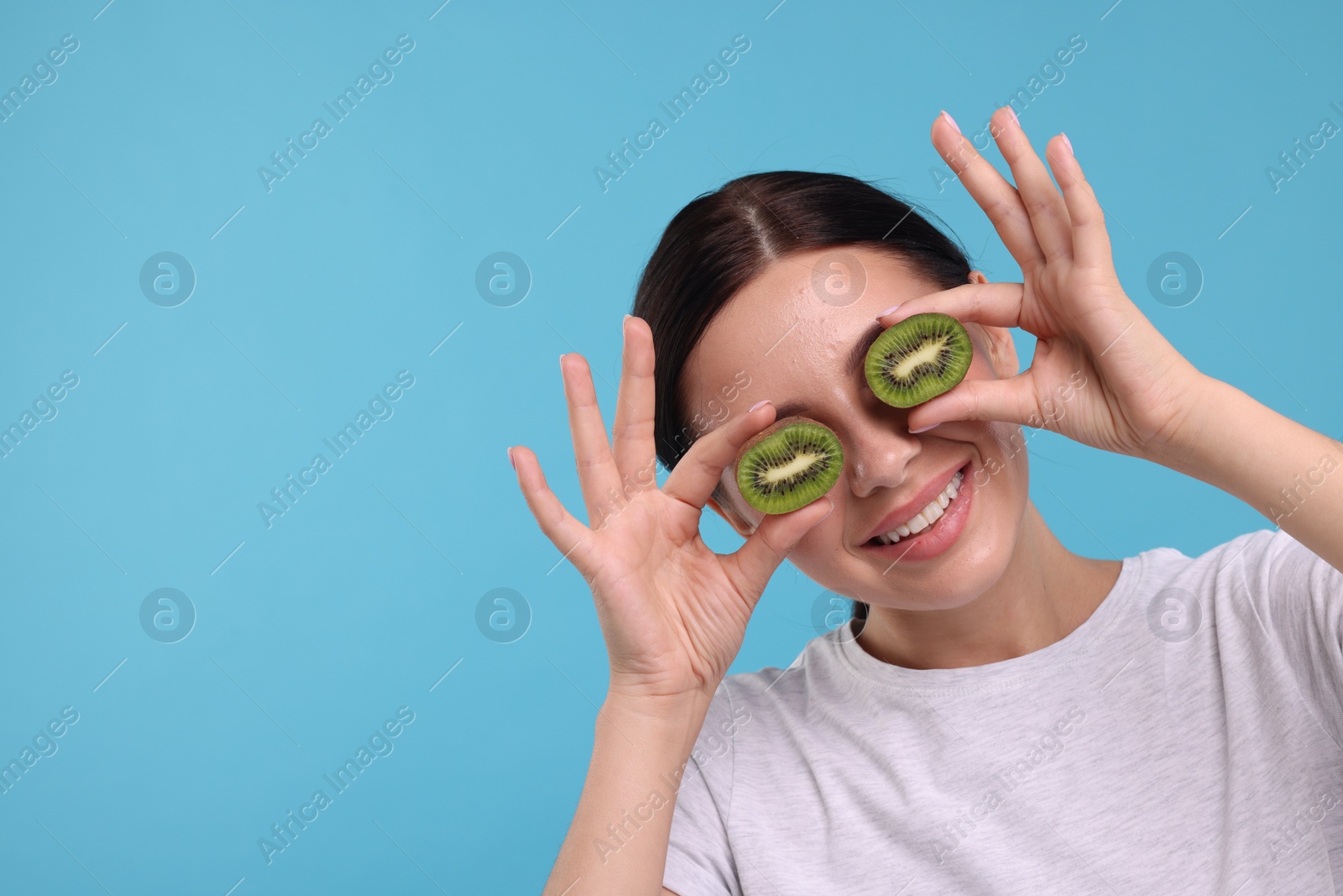 Photo of Young woman holding halves of kiwi near her eyes on light blue background, space for text