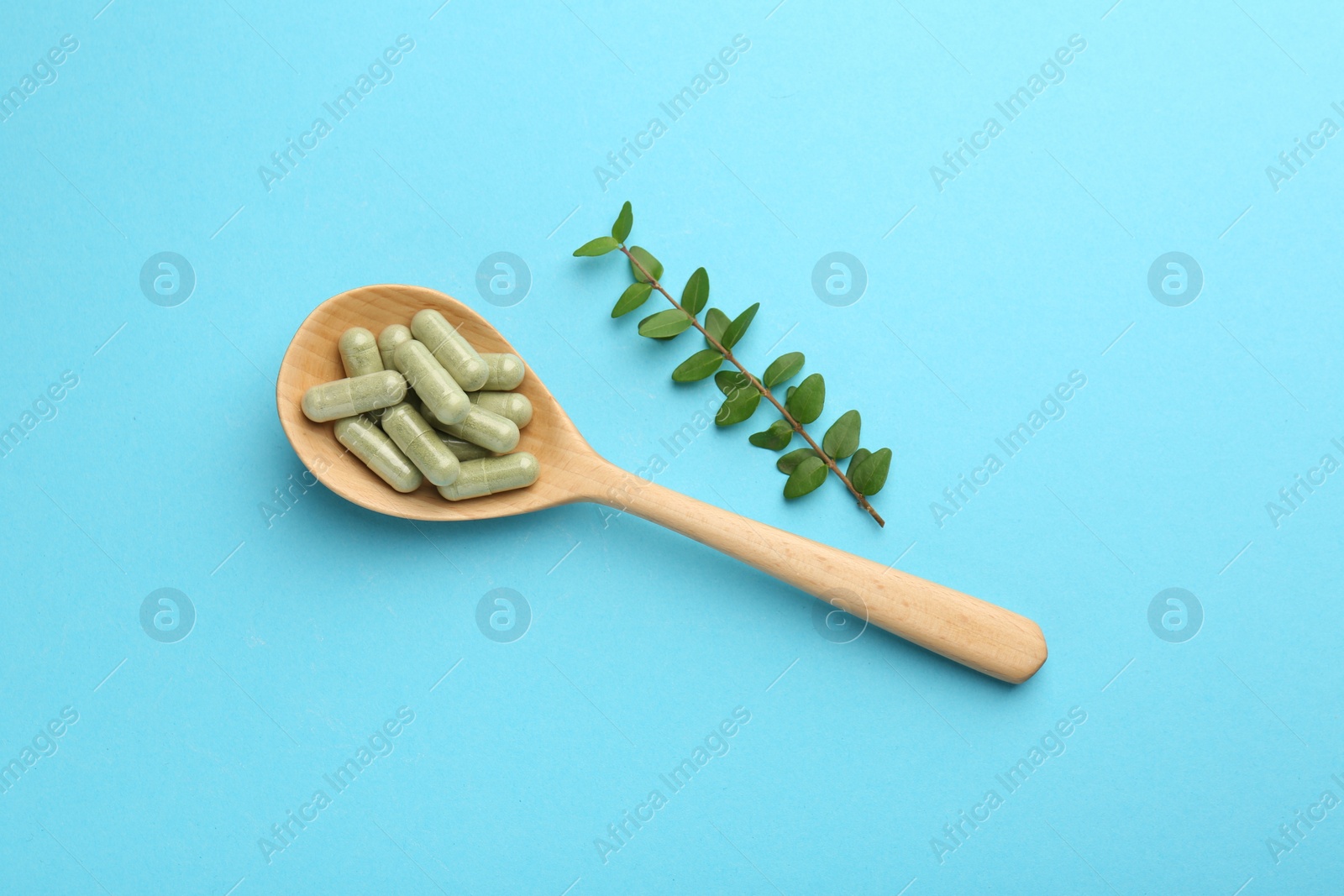 Photo of Vitamin capsules in spoon and green branch on light blue background, flat lay