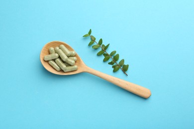 Vitamin capsules in spoon and green branch on light blue background, flat lay