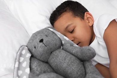 Photo of Cute little African-American boy with toy rabbit sleeping in bed