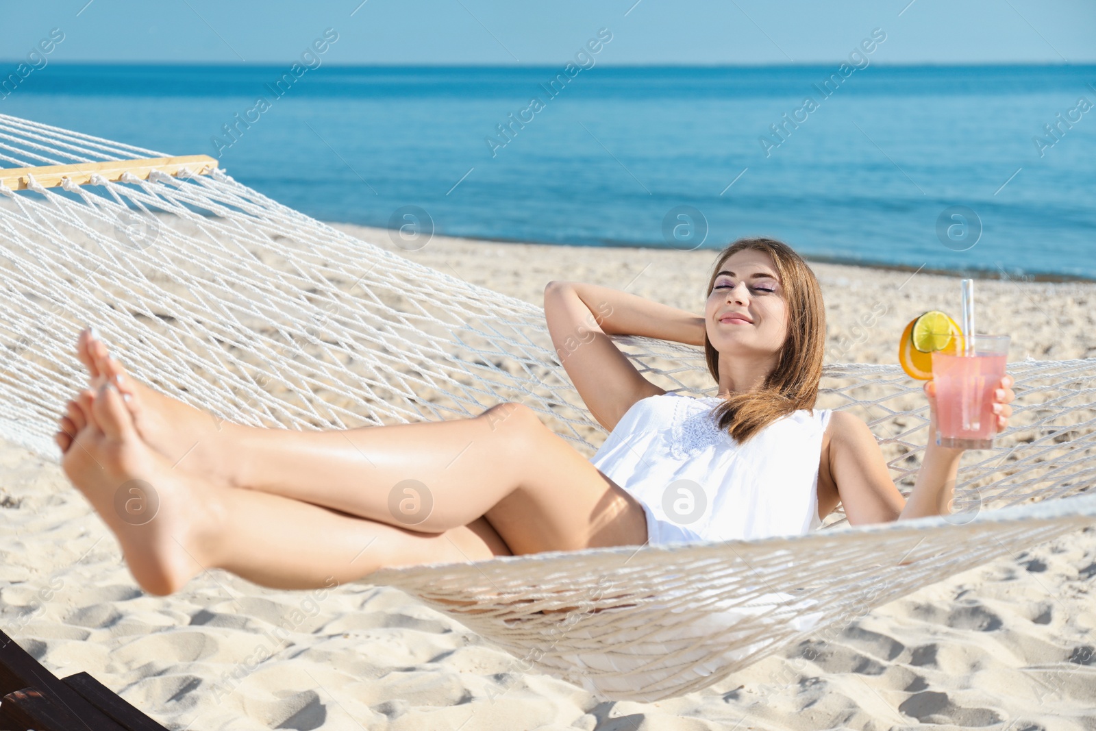 Photo of Young woman with refreshing cocktail relaxing in hammock on beach