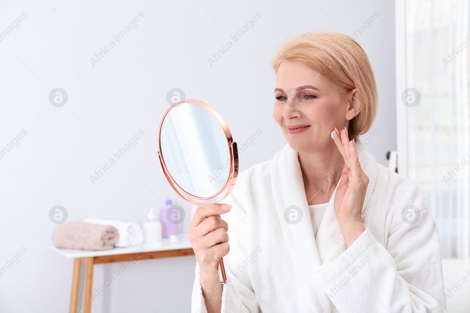 Photo of Mature woman with mirror applying face cream at home