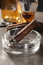 Photo of Smoldering cigar, ashtray and whiskey on grey table, closeup