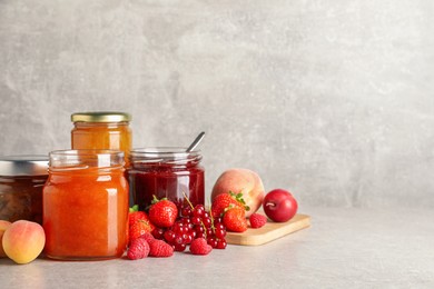 Jars with different jams and fresh fruits on grey table. Space for text