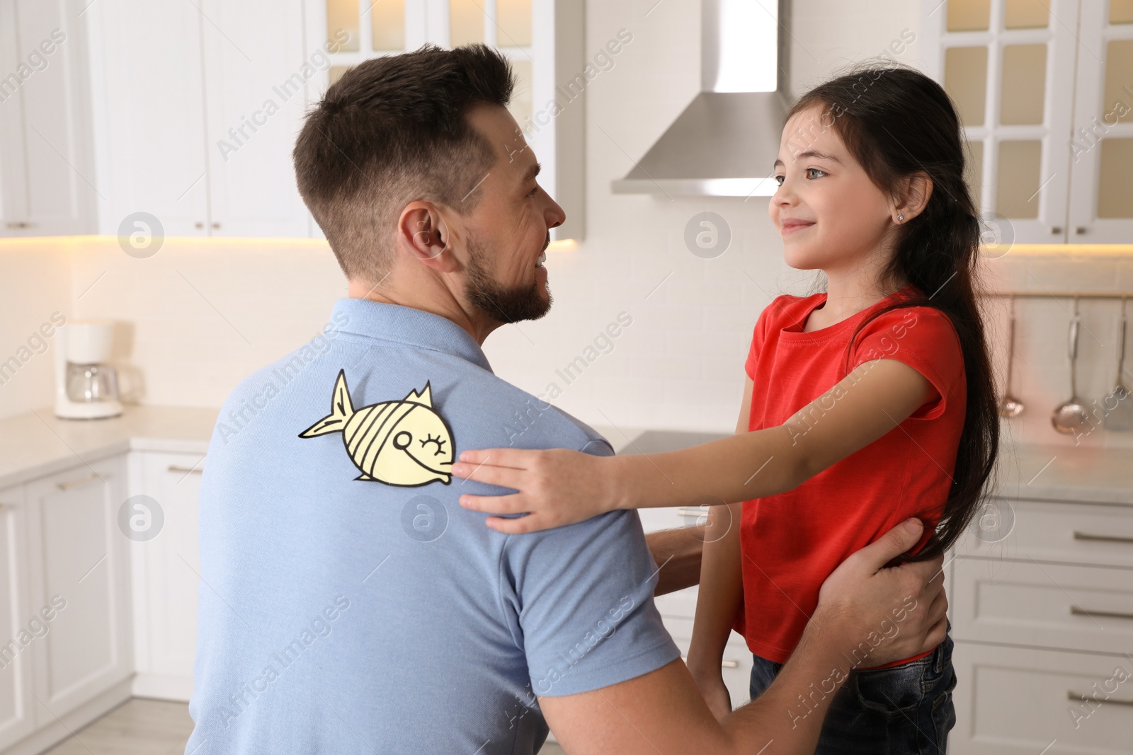 Photo of Cute little girl sticking paper fish to father's back at home