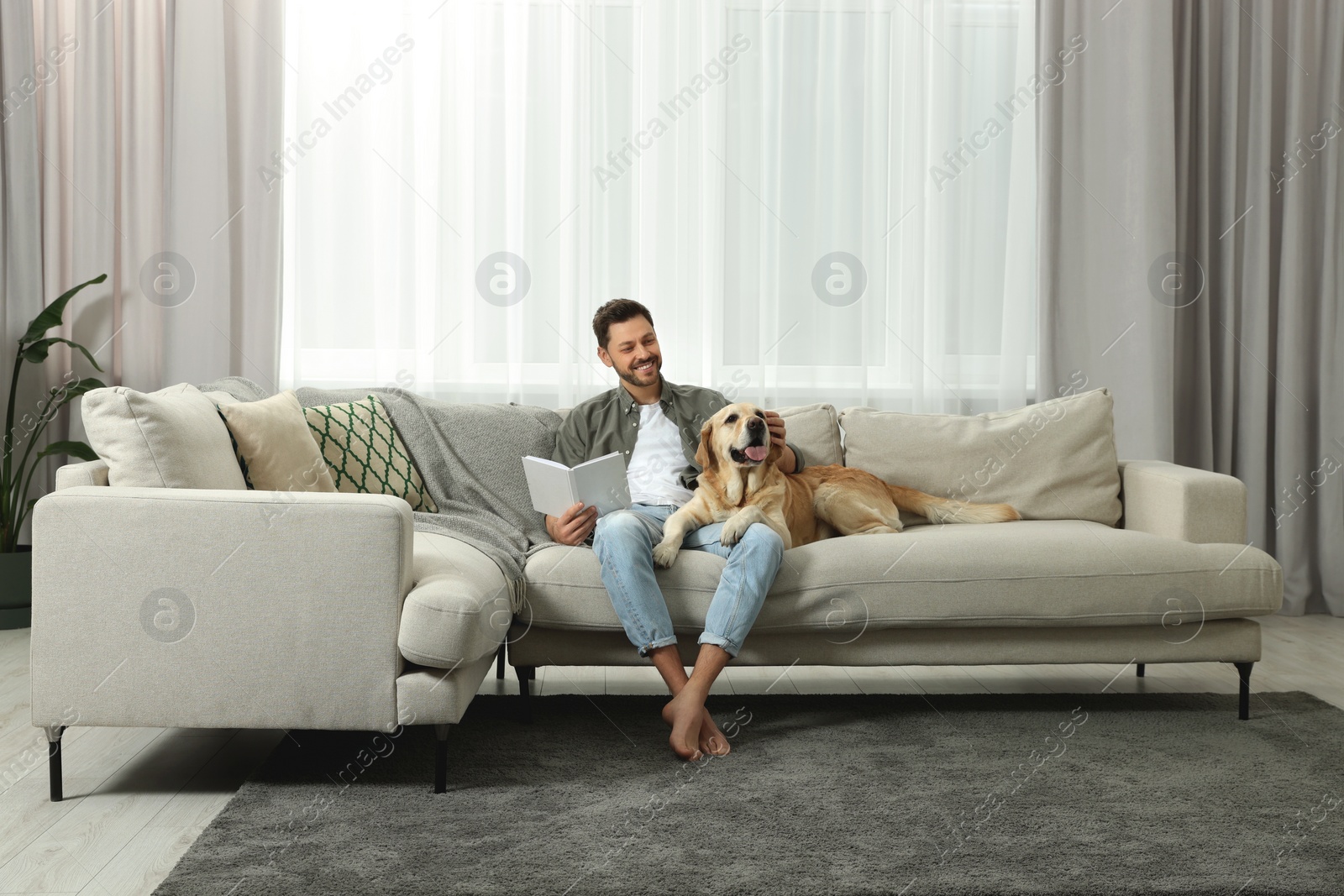 Photo of Man reading book on sofa near his cute Labrador Retriever at home