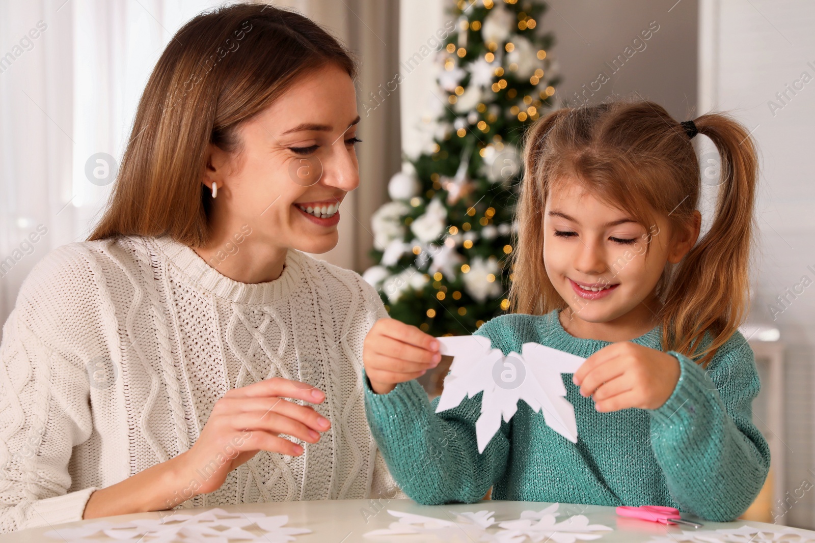 Photo of Happy mother and daughter making paper snowflake at table near Christmas tree indoors