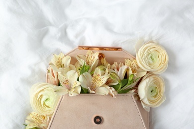 Photo of Stylish clutch with spring flowers on crumpled sheet, top view