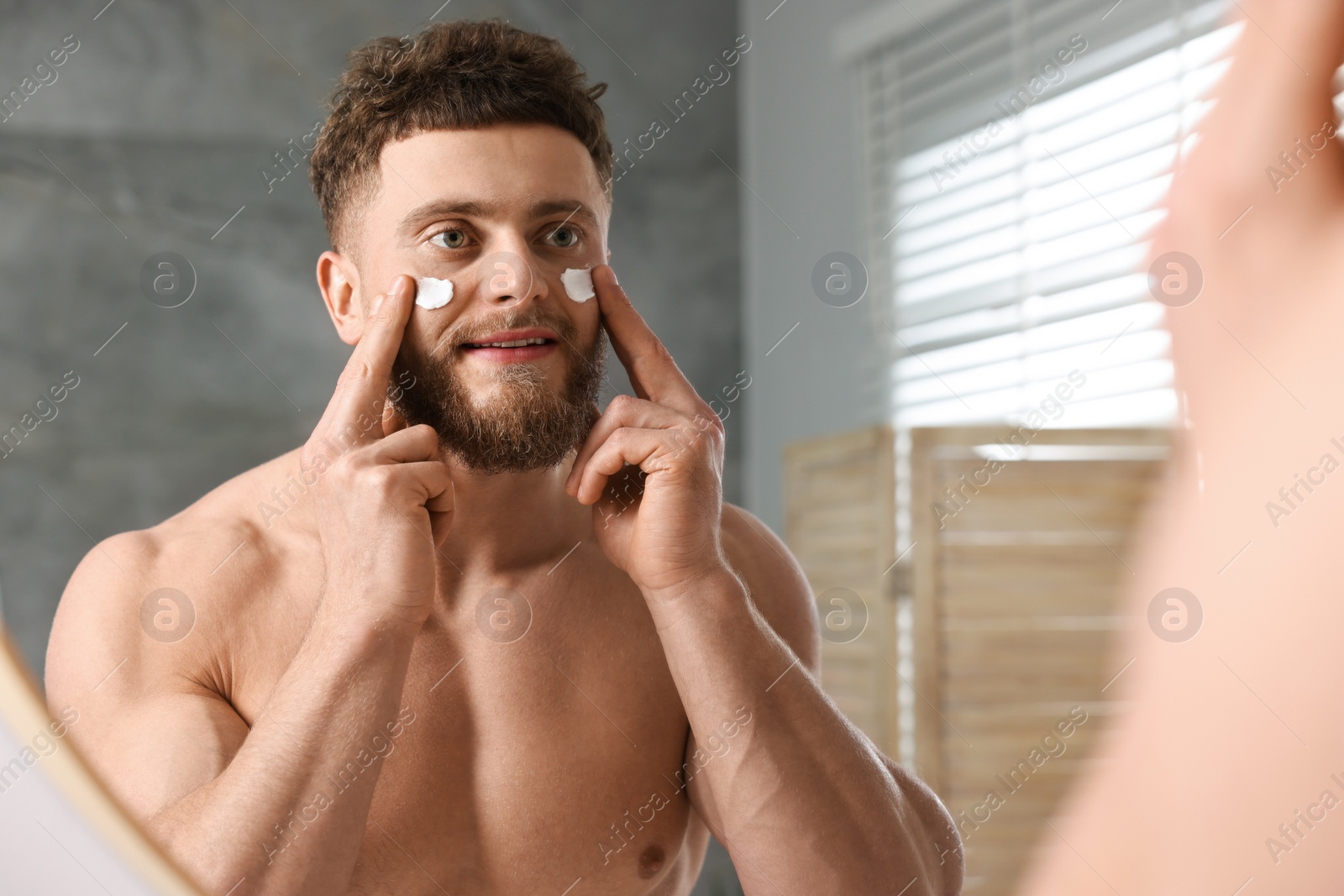 Photo of Handsome man applying moisturizing cream onto his face near mirror in bathroom