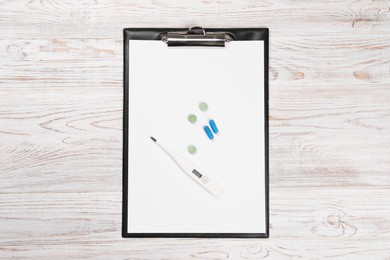 Clipboard, thermometer and pills on wooden table, top view