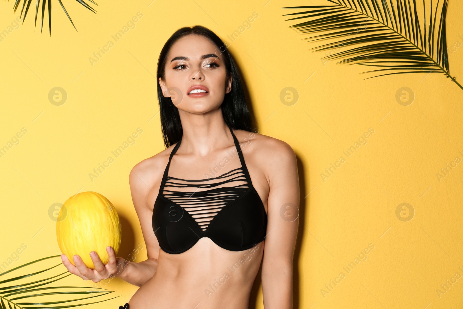 Photo of Beautiful young woman in black bikini with melon and tropical leaves on yellow background