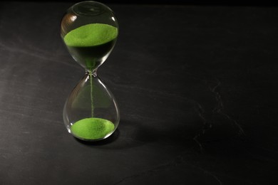 Photo of Hourglass with light green flowing sand on black table. Space for text