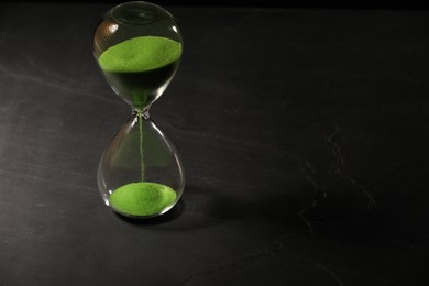 Hourglass with light green flowing sand on black table. Space for text