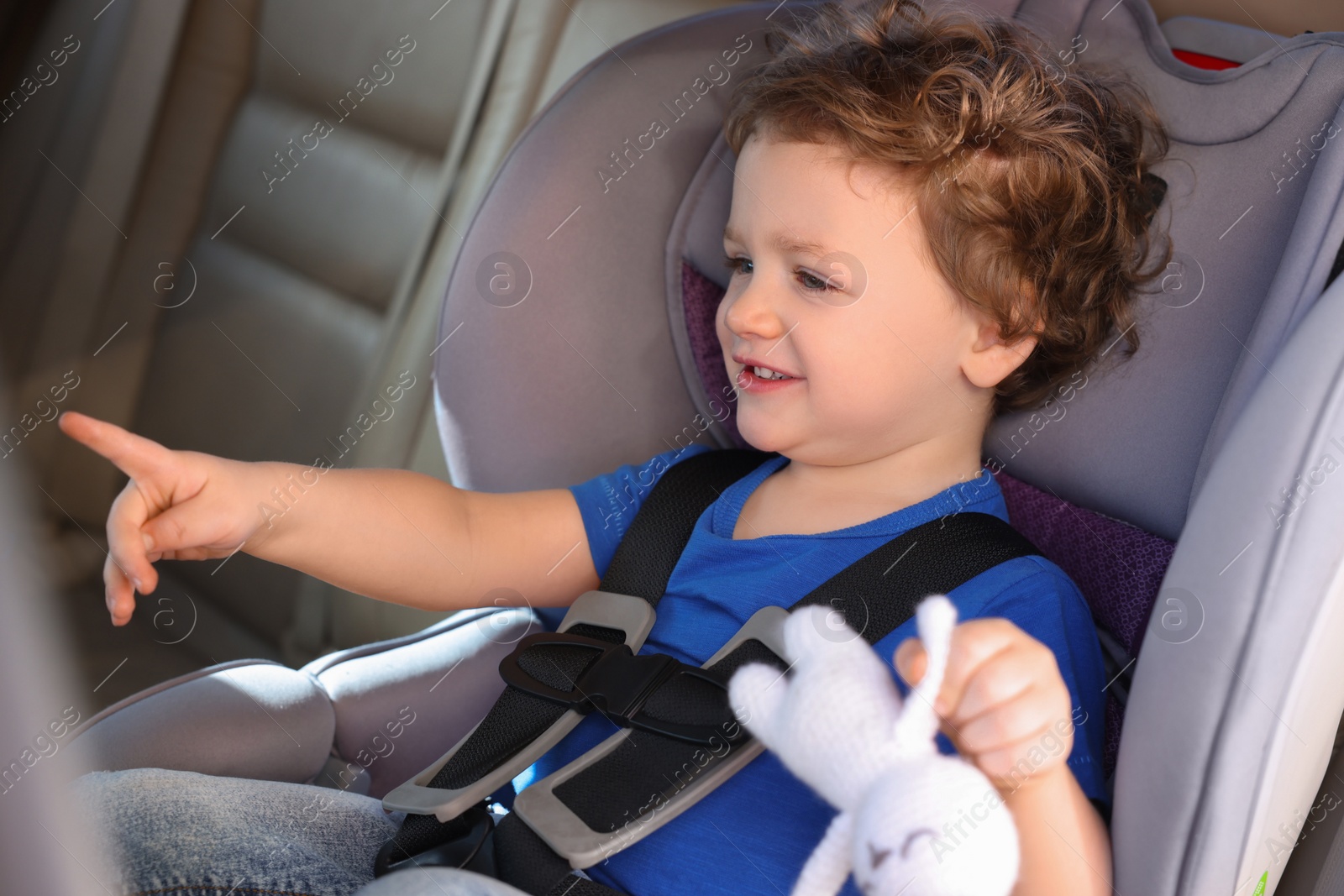 Photo of Cute little boy with toy rabbit pointing at something in child safety seat inside car