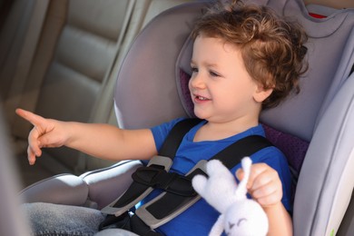 Photo of Cute little boy with toy rabbit pointing at something in child safety seat inside car