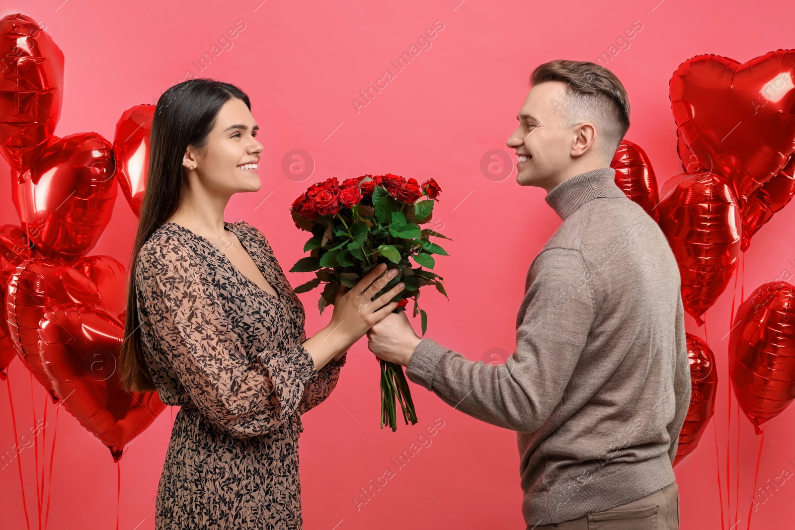 Photo of Boyfriend presenting bouquet of roses to his girlfriend near heart shaped air balloons on red background. Valentine's day celebration