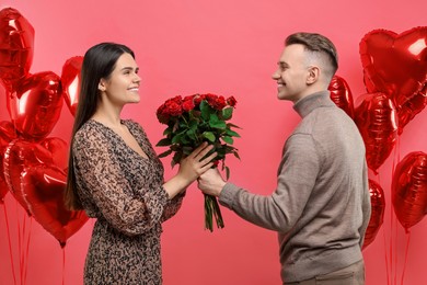 Photo of Boyfriend presenting bouquetroses to his girlfriend near heart shaped air balloons on red background. Valentine's day celebration