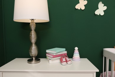 Photo of Books, booties, lamp and toy pyramid on chest of drawers in baby room